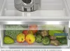 A close-up view of a refrigerator drawer filled with fresh fruits and vegetables, including apples, pears, broccoli, artichokes, and cabbage. A stainless steel pot is visible on the shelf above. Produktbild-Vorschau
