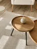 A close-up shot of a round wooden coffee table with a small vase of dried flowers on it, placed on a textured rug. Sunlight streams in from the side, casting shadows. Voir les images du produit