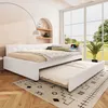 A white daybed with a pulled-out trundle bed is shown in a well-lit room with a large window overlooking a garden. The daybed has beige and patterned bedding and several decorative pillows. The room has a minimalist, modern design with natural wood floors  Podgląd zdjęcia produktu
