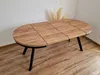 An oval dining table with a wood-grain top and black metal legs, set against a white wall and wooden floor. Produktbild-Vorschau
