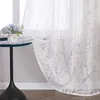 A close-up of a sheer curtain with a branch pattern hanging in front of a window. A small table with a vase of flowers is visible to the left. Produktbild-Vorschau