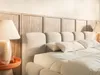 A close-up view of a bed with a light beige upholstered headboard and a wooden paneled wall behind it. Produktbild-Vorschau