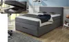 A bedroom scene featuring a box spring bed with storage, and a close-up inset showing the storage compartment open. Produktbild-Vorschau