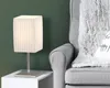 A table lamp sits on a white side table next to a gray armchair with a fluffy gray blanket draped over it. Produktbild-Vorschau
