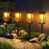 Four solar-powered garden lights are illuminating a pathway at dusk, surrounded by rocks and grass. Produktbild-Vorschau