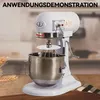 A white stand mixer with a silver bowl and whisk attachment is shown on a wooden surface, with wheat stalks to the left and a checkered cloth to the right. Produktbild-Vorschau