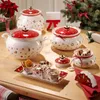 A collection of Christmas-themed ceramic dishes and bowls filled with cookies and dried oranges, arranged on a wooden table with a Christmas tree in the background. Anteprima dell’immagine prodotto