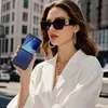 A woman in sunglasses and a white blazer holds a blue flip phone. Náhled obrázku produktu
