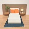 A mattress is displayed on a bed frame, with pillows and a bedside table with a lamp and books. Produktbild-Vorschau