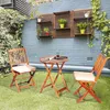 A small wooden bistro set with two chairs and a table is set up on a grassy patio area next to a brick wall with shelves holding plants. Náhľad obrázku produktu