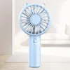 A light blue handheld fan with bear ears is shown in the center of the image, with a blurred background of a room with a sofa. Produktbild-Vorschau