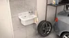A white sink with a faucet is mounted on a tiled wall in what appears to be a garage or utility room. A towel hangs from the faucet, and a car tire and part of a car are visible in the background. Anteprima dell’immagine prodotto