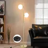 A modern floor lamp with two glass globes is placed next to a black leather sofa in a living room. Náhľad obrázku produktu