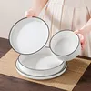 A person is holding a white plate with a black rim over a set of stacked plates and a bowl on a placemat. Voir les images du produit
