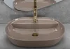 A beige oval sink with a gold faucet is shown in a modern bathroom setting with a concrete wall and mirror. Náhled obrázku produktu