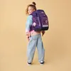 A young girl wearing a colorful sweater and baggy jeans, with a large purple backpack on her back, poses for a photo against a plain yellow background. Náhled obrázku produktu