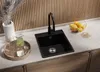 A black granite kitchen sink with a black faucet is shown on a countertop. Accessories like a pitcher of green liquid, glasses, and a bowl of fruit are also present. Produktbild-Vorschau
