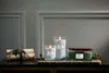 Three lit candles of varying sizes and colors are arranged on a decorative tray with some greenery and a decorative bundle of wood. Anteprima dell’immagine prodotto