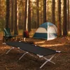 A camping scene in a forest with a cot in the foreground, a tent, and chairs in the background. Produktbild-Vorschau