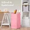 A pink felt laundry basket filled with rolls of craft paper is placed next to a white desk with a framed picture on it. A white shelving unit is visible on the right side of the image. Anteprima dell’immagine prodotto