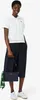 A woman is posing in a studio setting, wearing a white polo shirt and navy culottes, holding a navy handbag. Produktbild-Vorschau