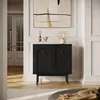 A black cabinet with arched doors stands in a well-lit room with wooden floors and white paneled walls. A large abstract painting hangs above the cabinet, and a table lamp sits on top. Produktbild-Vorschau