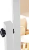 A close-up shot of a white stand mirror's adjustable knob and part of its frame. The background is blurred and shows a hint of a chair and patterned wallpaper. Náhled obrázku produktu