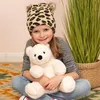 A young girl wearing a leopard-print hat with pom-poms sits on a rug, hugging a white teddy bear. A tall green plant in a basket is visible to her right. Produktbild-Vorschau