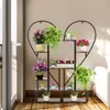 A heart-shaped plant stand filled with various potted plants is placed on a wooden floor near a balcony door and a white sofa. Produktbild-Vorschau