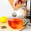A hand is holding a tea infuser over a glass mug filled with red tea. A lemon and loose tea leaves are on a wooden cutting board next to the mug. In the background, there is a jar of granola and a shelf. Voir les images du produit