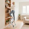 A woman is standing on a step stool next to a bookshelf, reaching for a book. Produktbild-Vorschau