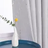 A white vase with a single yellow flower and green leaves sits on a blue tray in front of a gray curtain with silver stripes. Produktbild-Vorschau