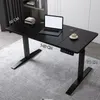 A black electric standing desk with a laptop, books, and a cup of coffee on it, placed on a grey carpet in a room with bookshelves in the background. Produktbild-Vorschau