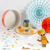 A birthday cake decorated with fondant circles and a small gift on top, placed on a cake stand, with party decorations and confetti in the background. Produktbild-Vorschau