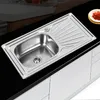 A stainless steel kitchen sink with a draining board is shown on a black countertop, with white cabinets below. Produktbild-Vorschau