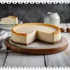 A cheesecake with a slice removed, placed on a wooden board, with a jug of milk and two nuts in the background. Produktbild-Vorschau