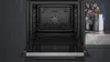 An open built-in oven with a view of the interior and a glimpse of a shelf with decorative items in the background. Produktbild-Vorschau