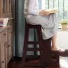 A person is sitting on a wooden step stool and reading a book next to a window. Náhľad obrázku produktu