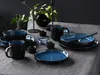 A table setting with blue and black stoneware plates, bowls, and mugs, arranged on a gray tablecloth. Produktbild-Vorschau
