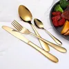 A set of gold-colored cutlery is arranged on a white surface, with a blurred background showing a plate of food. Anteprima dell’immagine prodotto