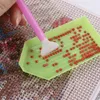 A close-up of a person's hand using a pink diamond painting tool to pick up red diamonds from a green tray, with a diamond painting canvas in the background. Produktbild-Vorschau