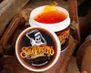 A jar of Suavecito pomade is shown open, with some product on a brush, placed on a surface of rusty metal objects. Podgląd zdjęcia produktu