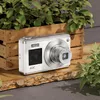 A white digital camera is placed on a wooden plank, with a wooden crate and plants in the background. Produktbild-Vorschau
