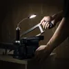 A person is using a hacksaw to cut a metal object clamped in a vise, illuminated by a flexible LED work light. Produktbild-Vorschau