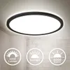 A modern circular ceiling light fixture with a black frame and a white illuminated diffuser, shown mounted on a ceiling with a brick wall in the background. Circular icons with text detailing the product's features are displayed in the foreground. Produktbild-Vorschau