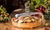 A glass cake stand with a pie covered by a glass dome, set on a wooden table with fresh vegetables and herbs in the background. Náhľad obrázku produktu