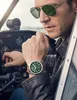 A man wearing sunglasses and a leather jacket sits in the cockpit of a small airplane, looking at his watch. Náhľad obrázku produktu