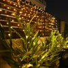 A string of warm white fairy lights is draped over a wooden fence and plants at night. Náhled obrázku produktu