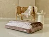 A pet bed is placed on the floor in front of a rattan chair with a blanket draped over it. A small white side table with a candle is next to the chair. Produktbild-Vorschau