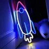 A neon rocket sign is displayed in a room, casting colorful light on the wall and floor. A stuffed animal is visible in the background. Produktbild-Vorschau